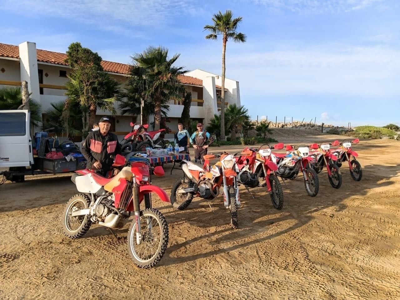 Motorcyclists lined up on dirt beside a white building, with palm trees and blue sky in the background