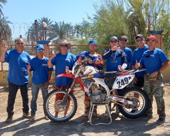 Group of people in blue shirts posing with a dirt bike in a sunny outdoor desert setting