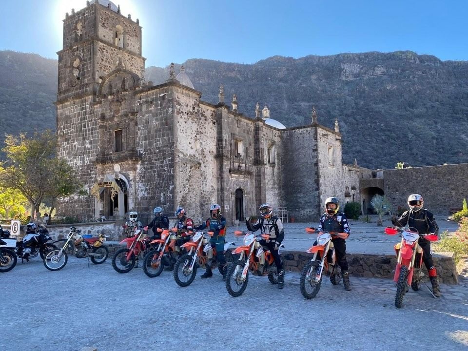 Motorcycles parked outside a stone church with mountains in the background