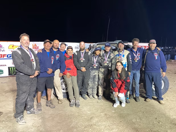 Group of people posing at night on a dirt track, some in racing suits, with a race car in the background