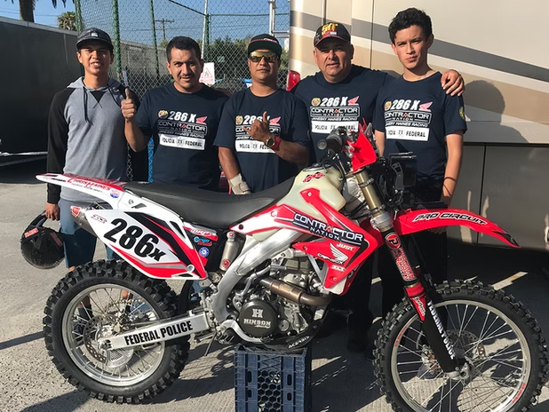 Five men posing behind two motocross bikes, including a red bike and a white bike with race number 286X.