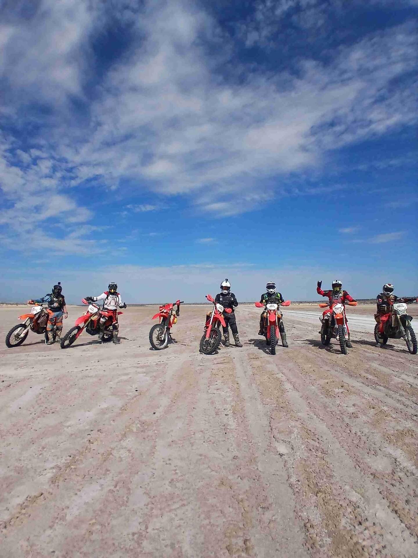 Six motocross riders lined up on a dusty desert road under a bright blue sky.