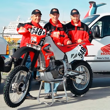 Three riders in red Honda gear posing with a red and white motocross bike beside a Honda van at a track.