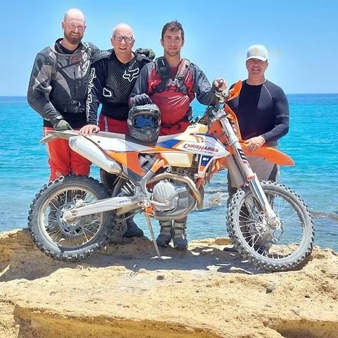 Four dirt bikers posing with a KTM motorcycle on a sandy beach beside turquoise water.