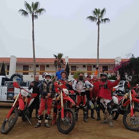 Group of motocross riders posing with red dirt bikes in front of a building and palm trees.