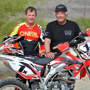 Two men pose with a red and black dirt bike, both in racing gear and smiling outdoors.