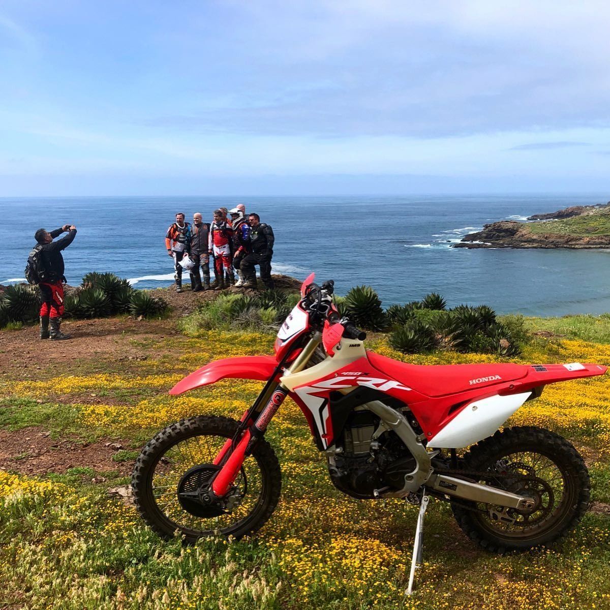 Red and white dirt bike parked by a coastal overlook with a group of people in the background.