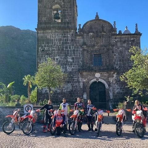 Group of motorcyclists on dirt bikes posing in front of a historic stone church in sunlight