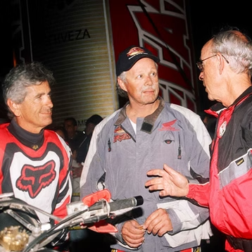 Three men in motorcycle gear talking beside a bike at night.