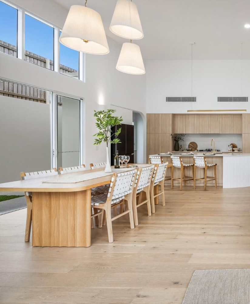 Bright modern dining room with long wooden table, white chairs, pendant lights, and open kitchen. 
