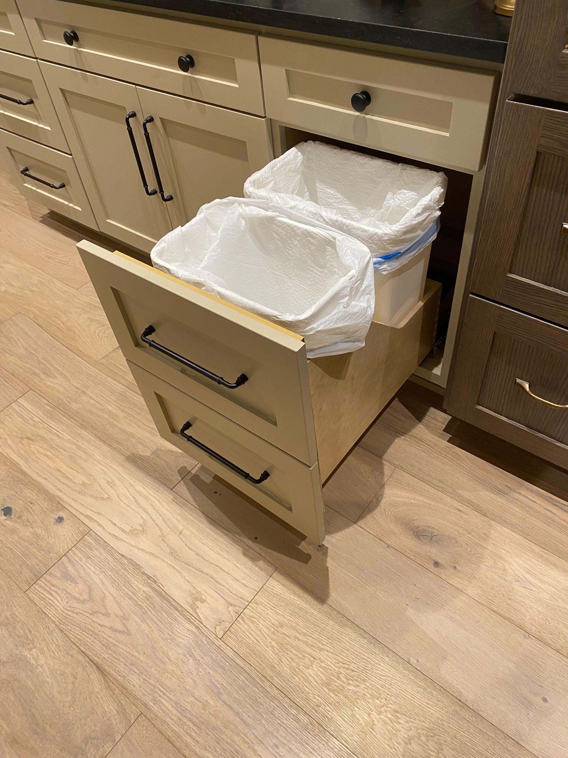 Open kitchen trash drawer with a white trash liner pulled out and hanging over the edge.