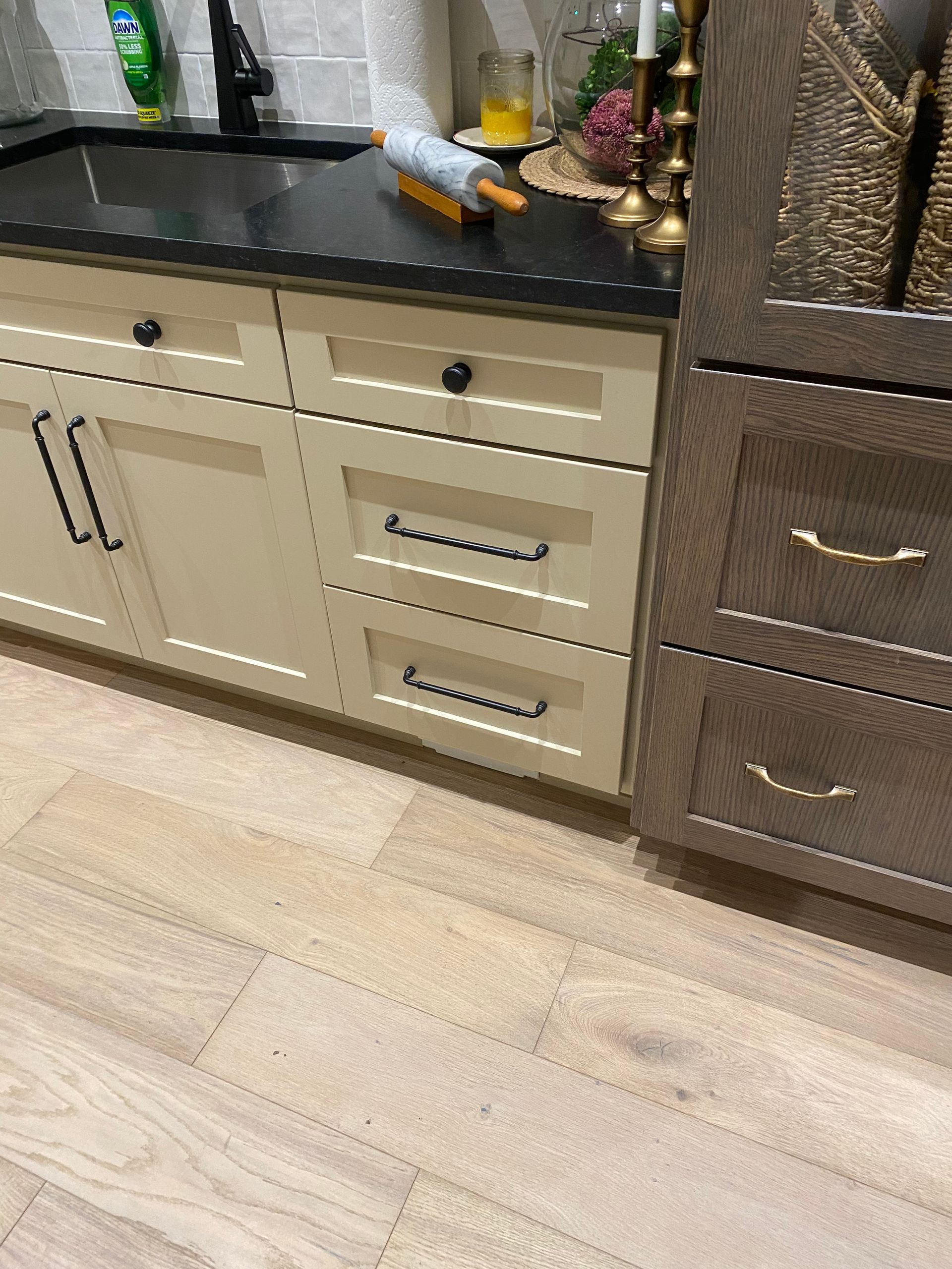 Kitchen cabinets with black countertop, beige drawers, and wood flooring.