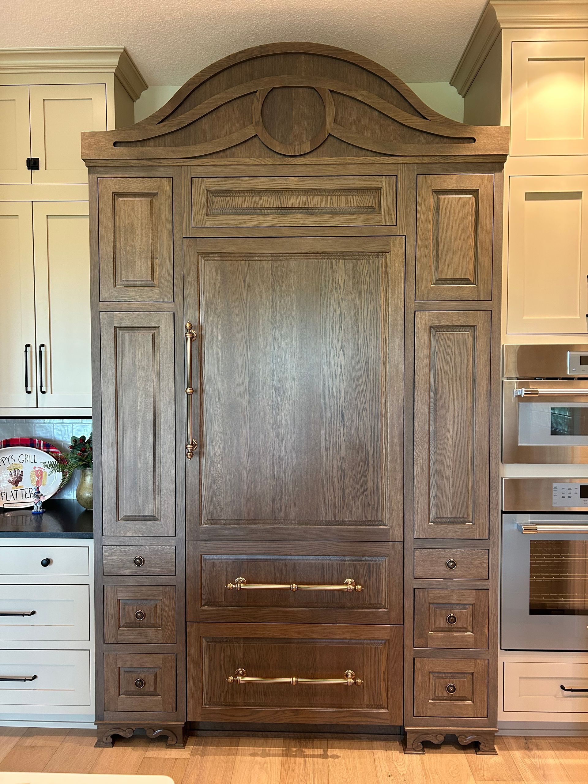 Ornate dark wood pantry cabinet built into a kitchen between white cabinets and stainless steel appliances