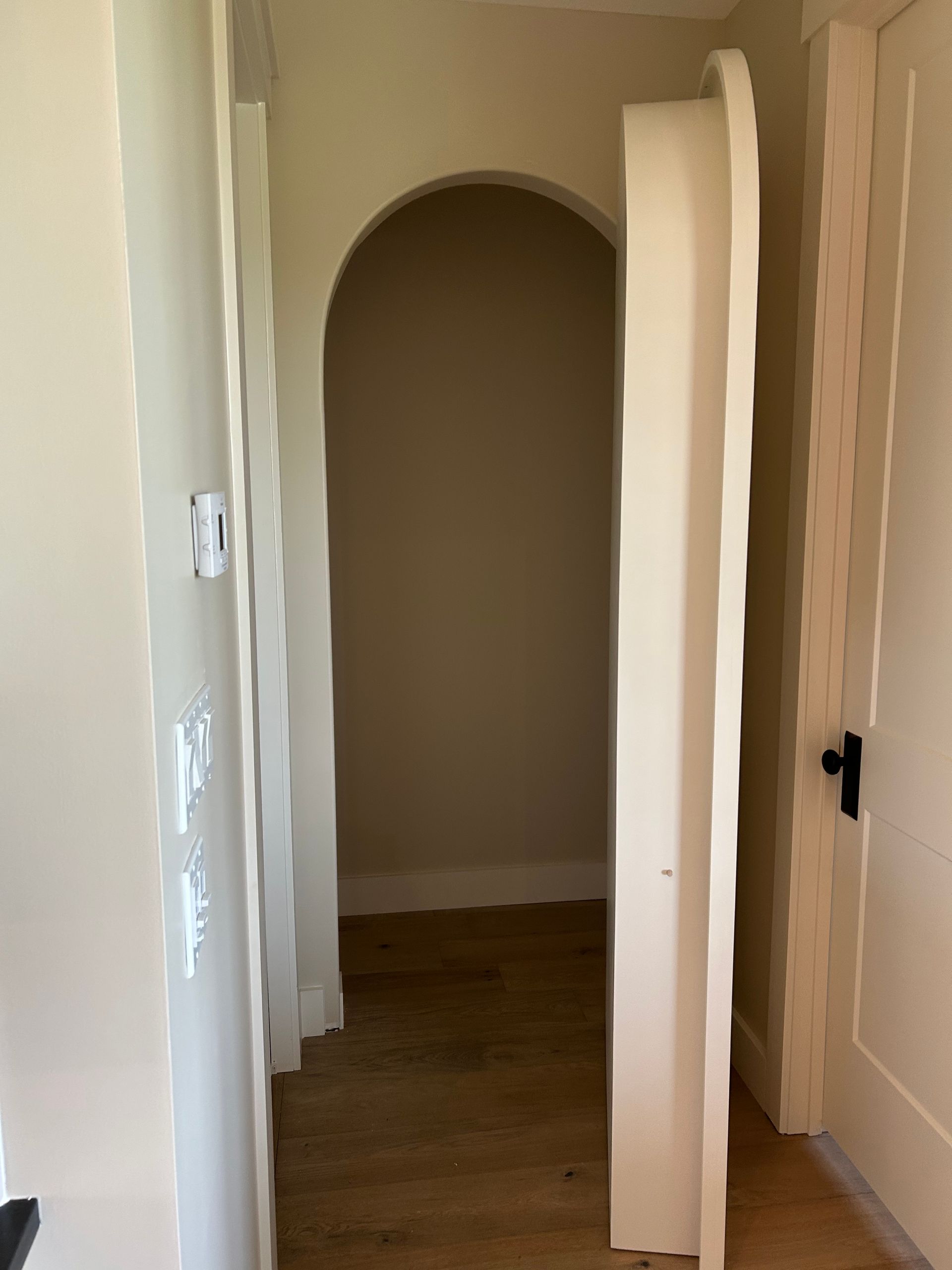 Narrow hallway with arched doorway, white walls, and wood flooring.