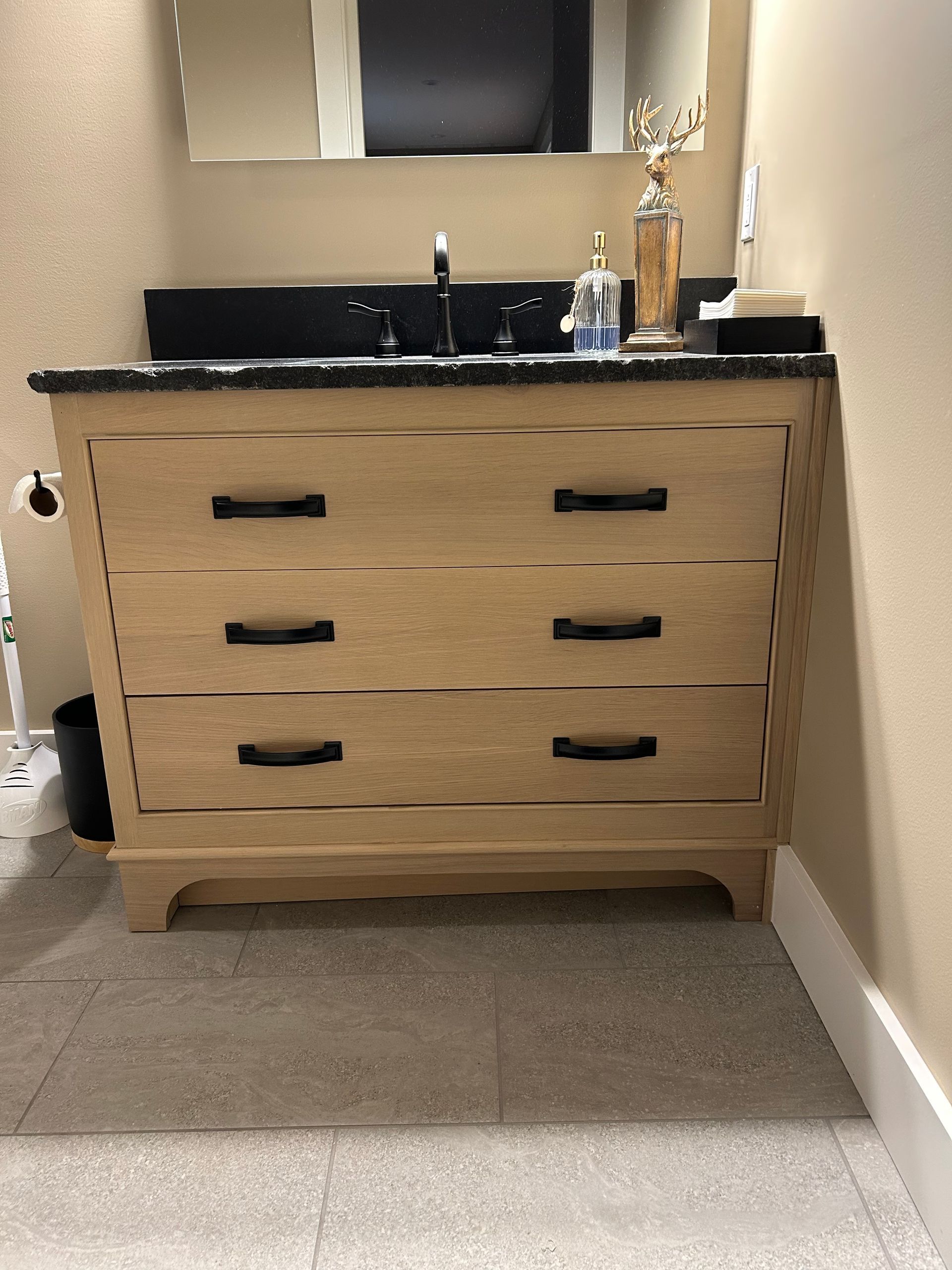 Wooden vanity with black countertop and mirror in a narrow hallway, with a sink and decor on top