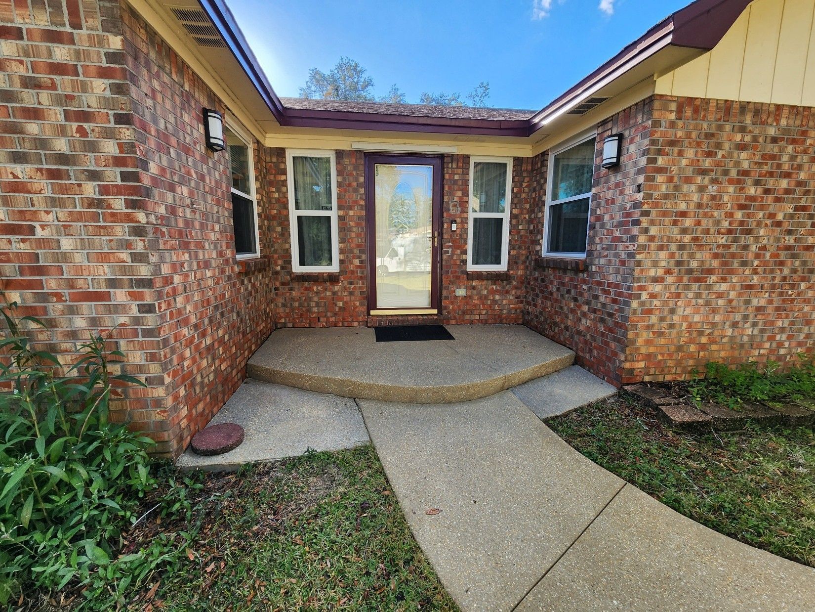 Front entry walkway to a brick house with a glass door, narrow windows, and a curved concrete path. Front entry walkway to a brick house with a glass door, narrow windows, and a curved concrete path.