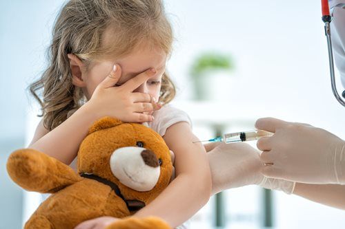 Child getting a vaccine shot from a healthcare professional, covering face with one hand, holding a teddy bear.