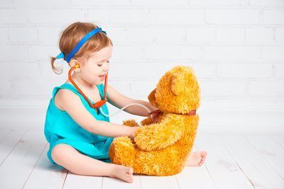 Young child in blue dress playing doctor with teddy bear, using stethoscope.