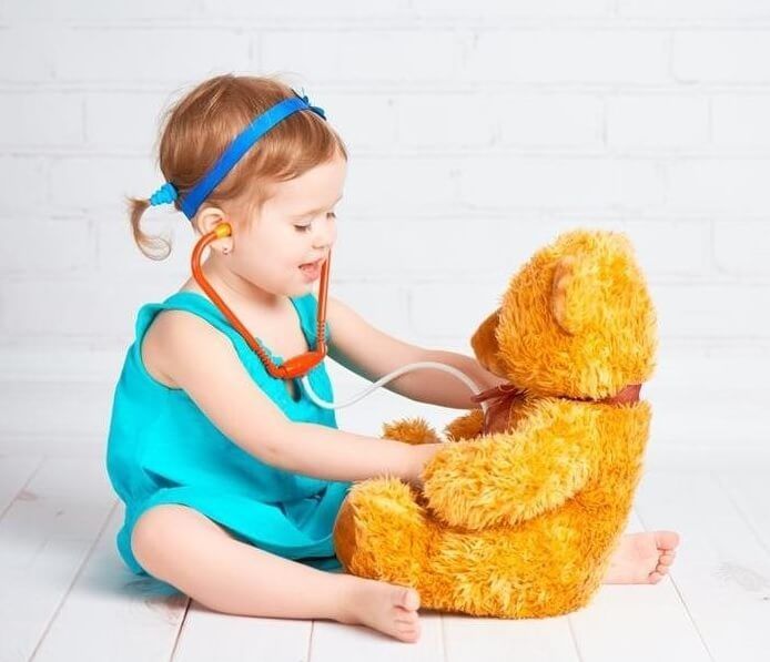 Young child in blue dress, playing doctor with a teddy bear, using a stethoscope.