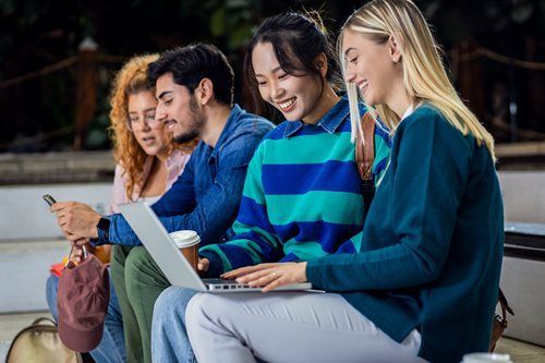 Four students looking at laptop outdoors; smiling.