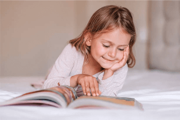 Girl smiles while reading a book, lying on a white bed.