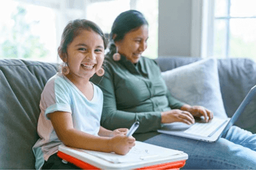 Girl smiling, holding pen, seated next to woman using laptop on couch.