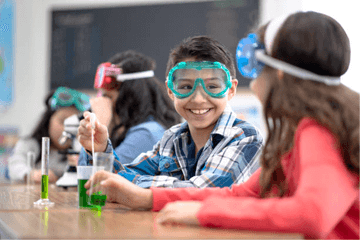 Kids in science class, mixing green liquid, wearing goggles, smiling.
