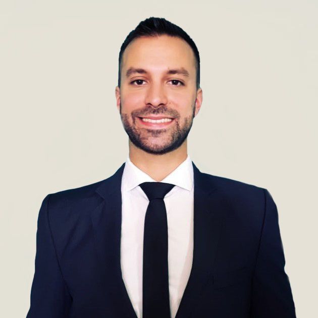 Hombre sonriente con traje oscuro y corbata sobre un fondo claro liso.