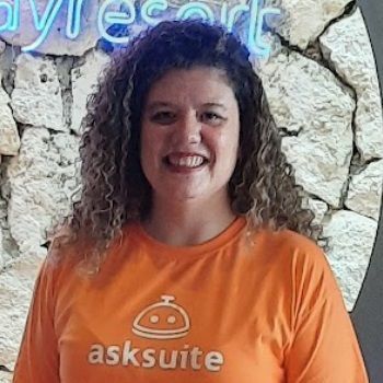 Mujer con una camiseta naranja de Asksuite sonriendo frente a un muro de piedra y un letrero de neón.
