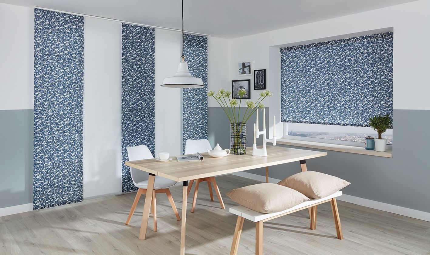 Dining room with a light wood table, bench, and chairs. Blue patterned panels and window shades, white walls. Abundance Blinds In Charmhaven, NSW