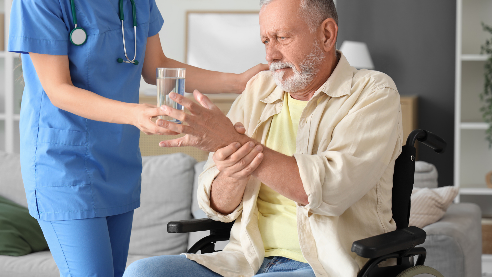 Nurse in blue helping a seated patient drink water in a bright living room