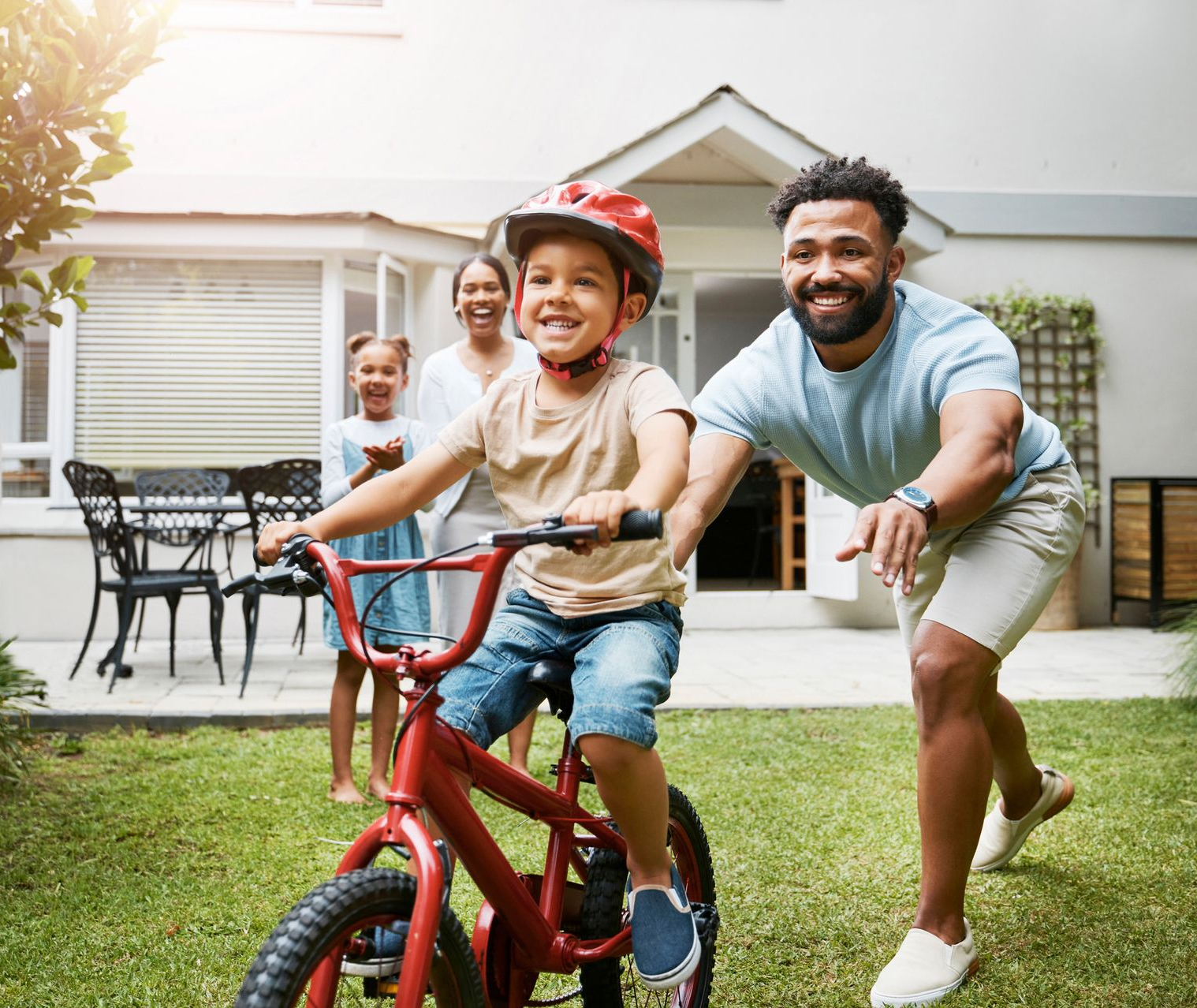 A father helps his child learn to ride a bike in a backyard. Family watches.