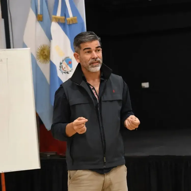 Un hombre habla frente a una bandera argentina, lleva un chaleco negro y gesticula con ambas manos.