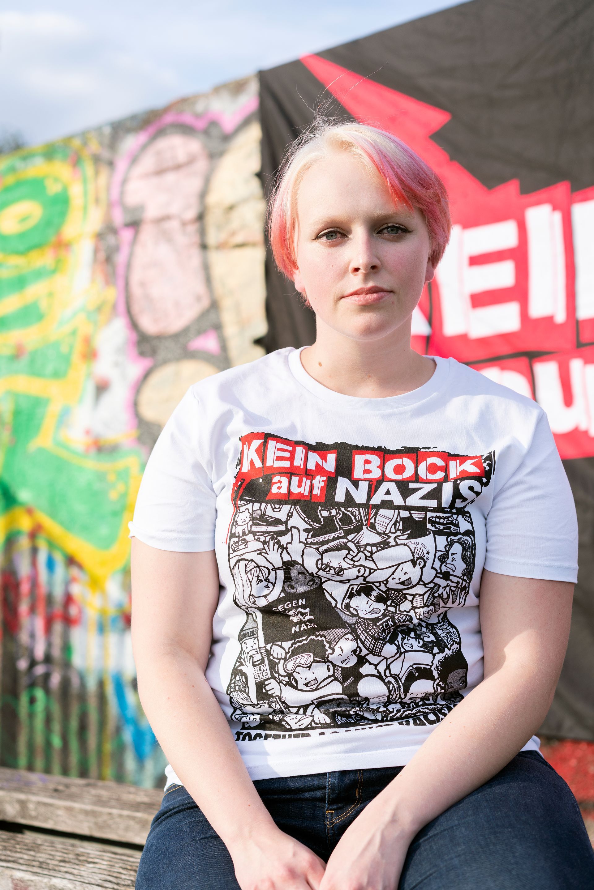 Eine Person mit pinkfarbenen Haaren und einem T-Shirt mit der Aufschrift „Kein Bock auf Nazis“ sitzt vor Graffiti.