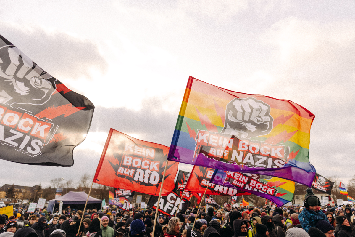 Eine Menschenmenge protestiert mit Fahnen mit der Aufschrift „Keine Nazis“ und einer Regenbogenfahne.