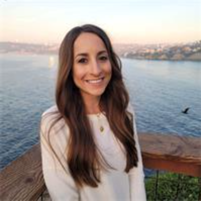 Smiling woman in white top standing by a waterfront at sunset with cityscape in the background