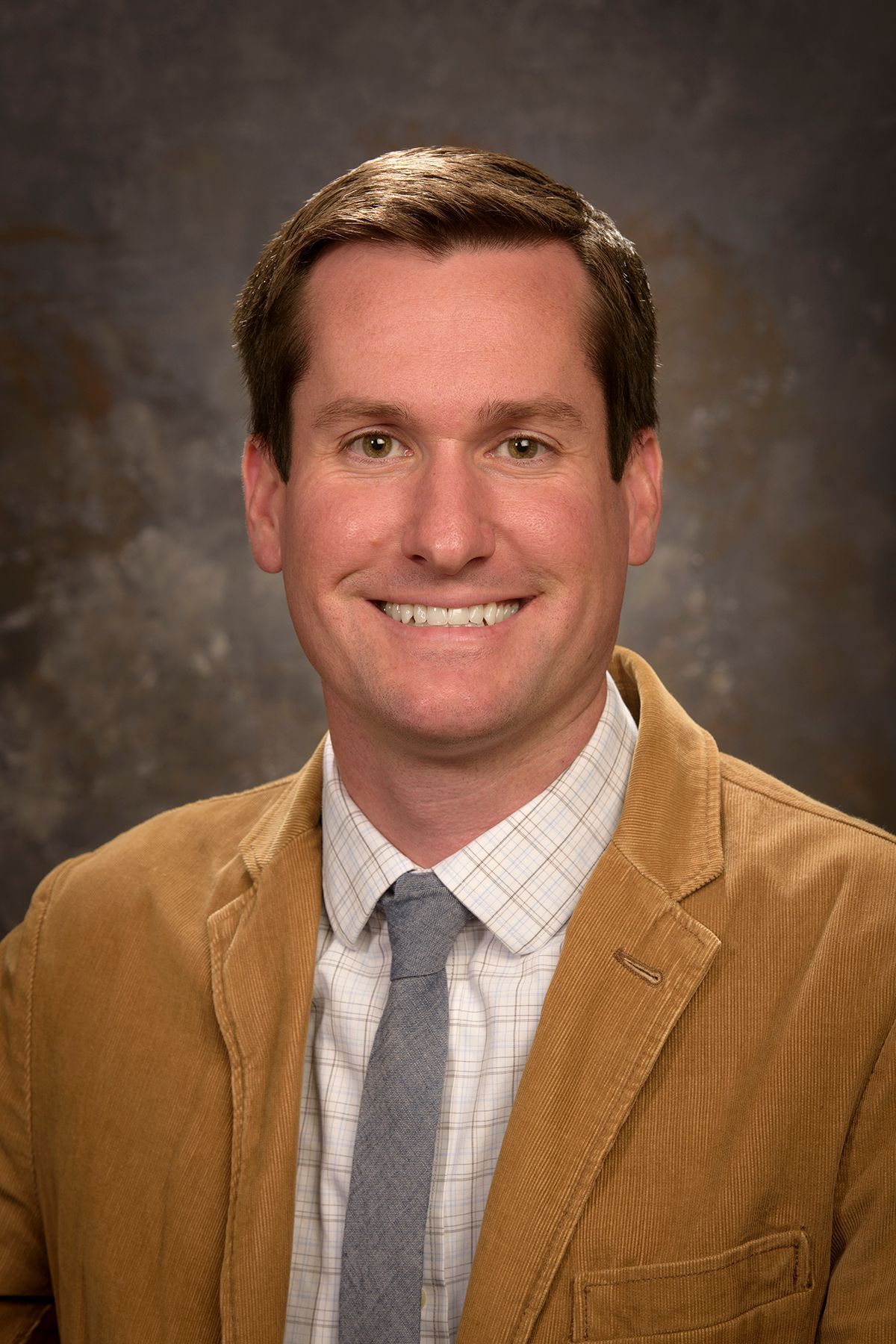 Portrait of a smiling man in a tan jacket, gray tie, and white shirt against a studio backdrop