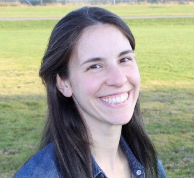 Smiling woman with dark hair in a blue jacket outdoors on a grassy field