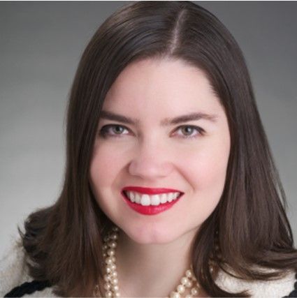 Smiling woman with dark hair, red lipstick, and pearl necklace against a gray background