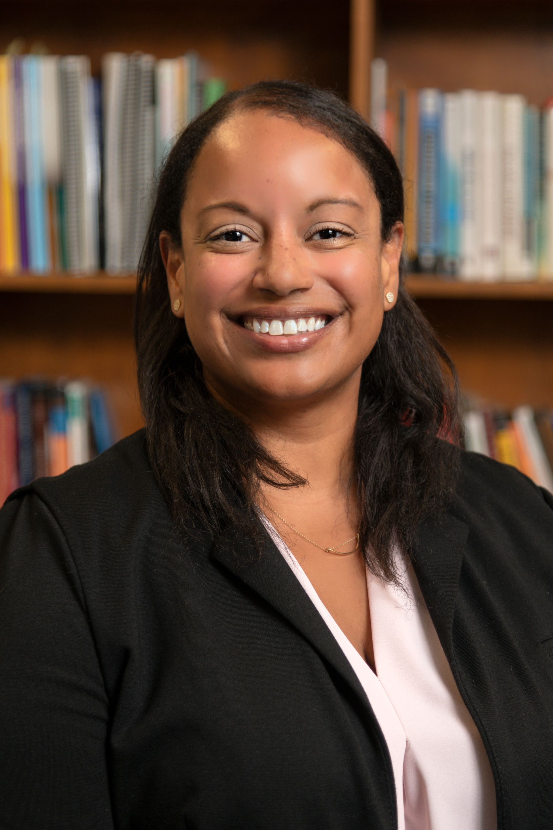 Smiling person in a black blazer standing before a bookshelf
