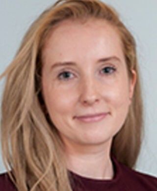 Portrait of a smiling person with blonde hair in a maroon top against a light gray background