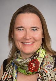 Smiling woman in a patterned scarf and brown top against a gray background