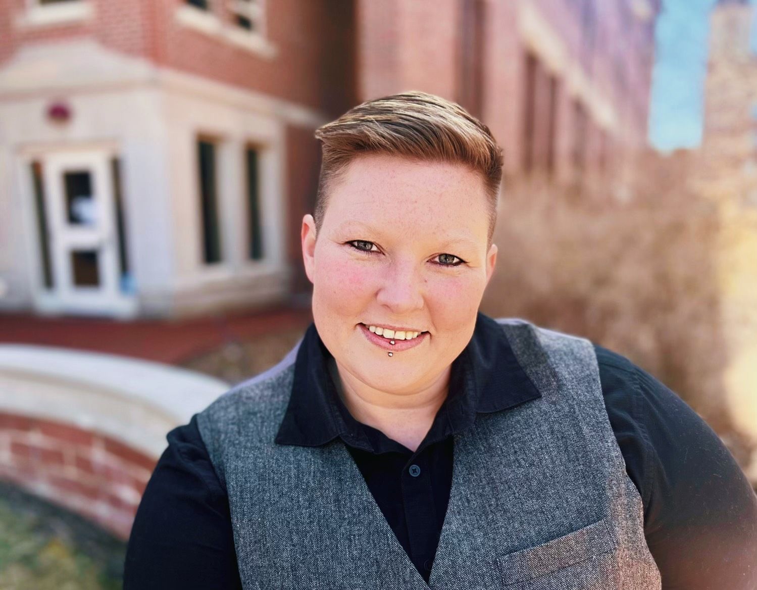 Smiling person in a gray vest standing outdoors near a brick building