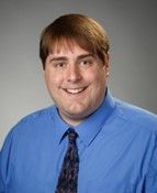 Portrait of a smiling person in a blue shirt and patterned tie on a gray background