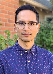 Portrait of a man wearing glasses and a navy patterned shirt, standing outdoors near a brick wall and greenery