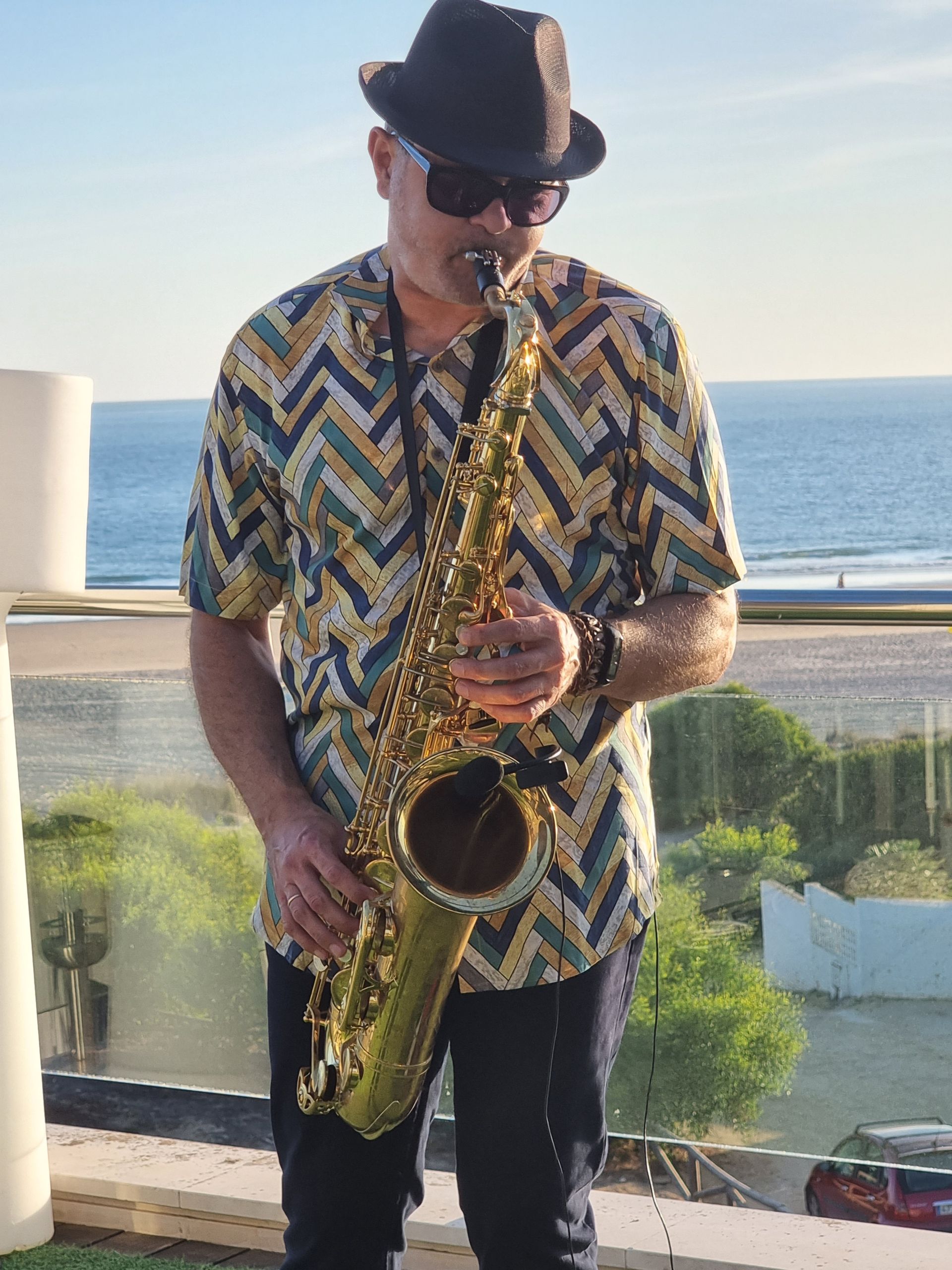 Hombre tocando el saxofón al aire libre; el océano al fondo. Lleva sombrero, gafas de sol y camisa estampada.