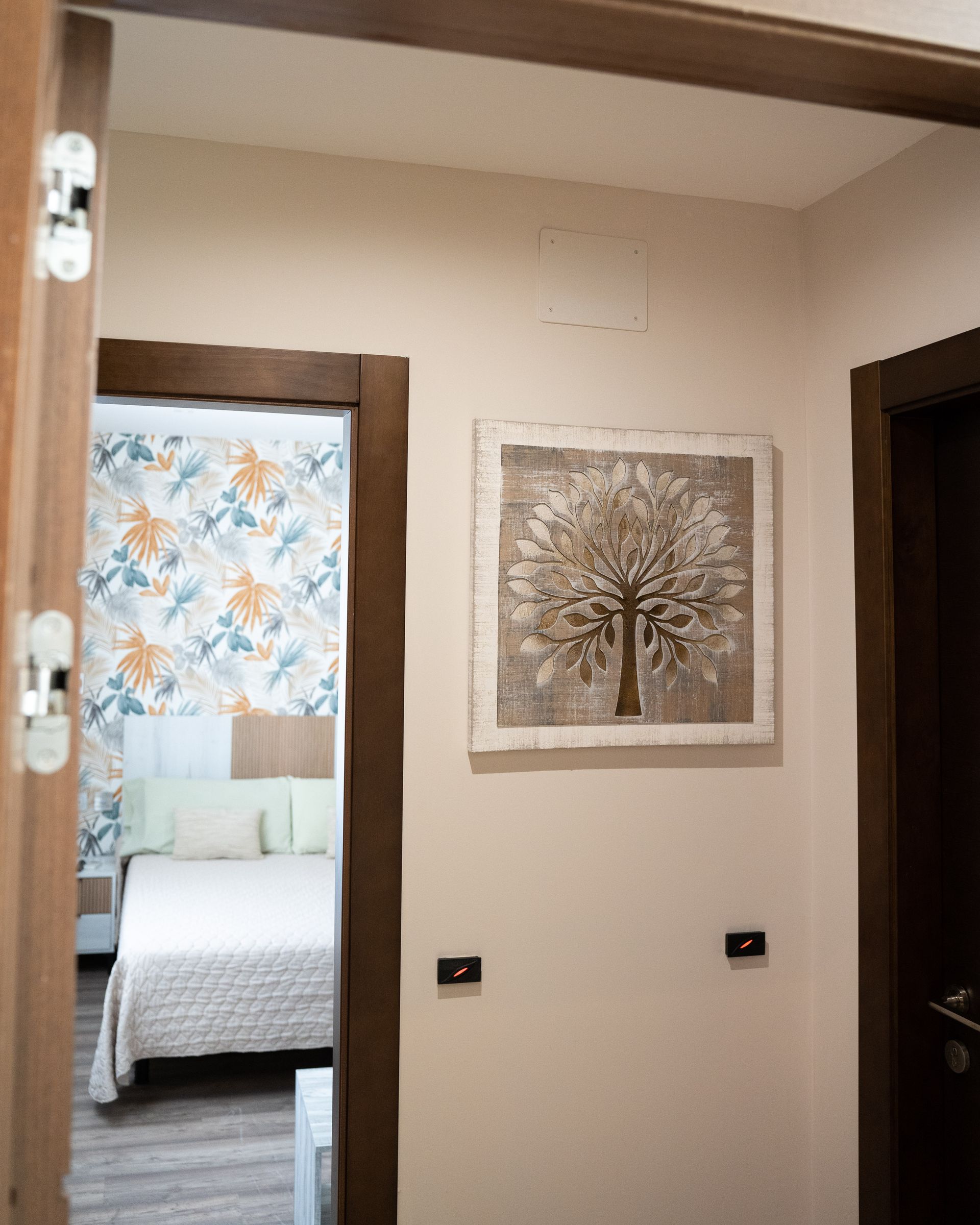 Pasillo con una pieza de arte de árbol; dormitorio con papel tapiz floral y una cama visible.