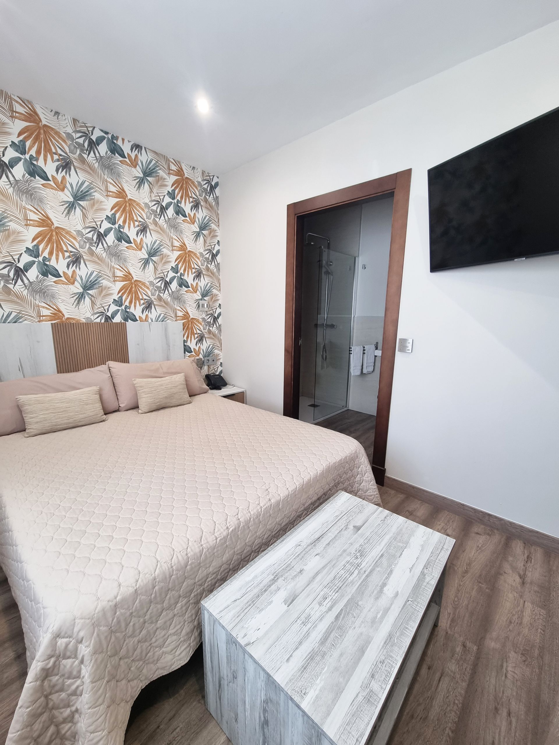 Dormitorio con cama, papel pintado estampado, mesa pequeña y baño a la vista.