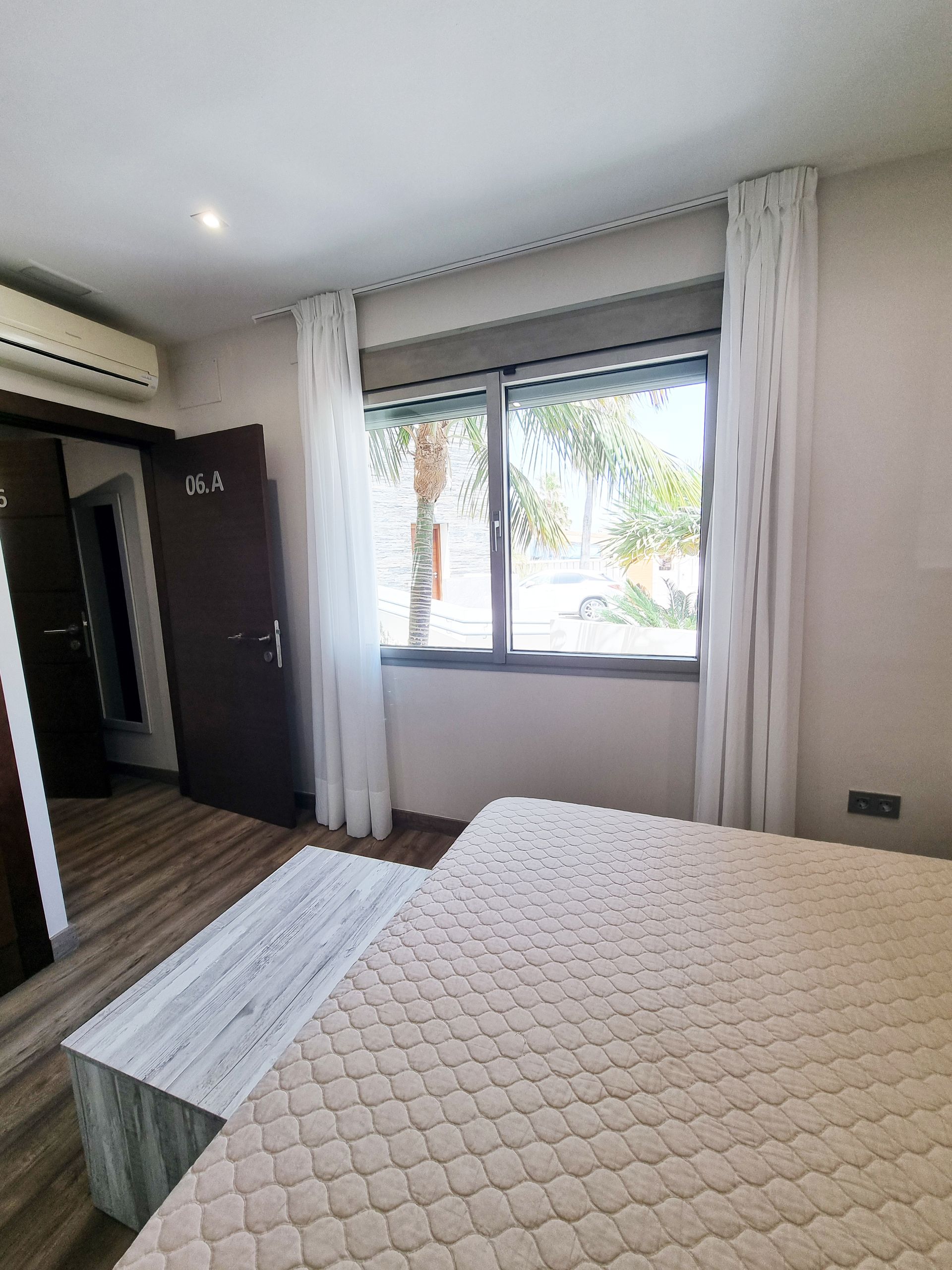 Dormitorio con cama, banco, ventana con cortinas transparentes, armario y aire acondicionado.