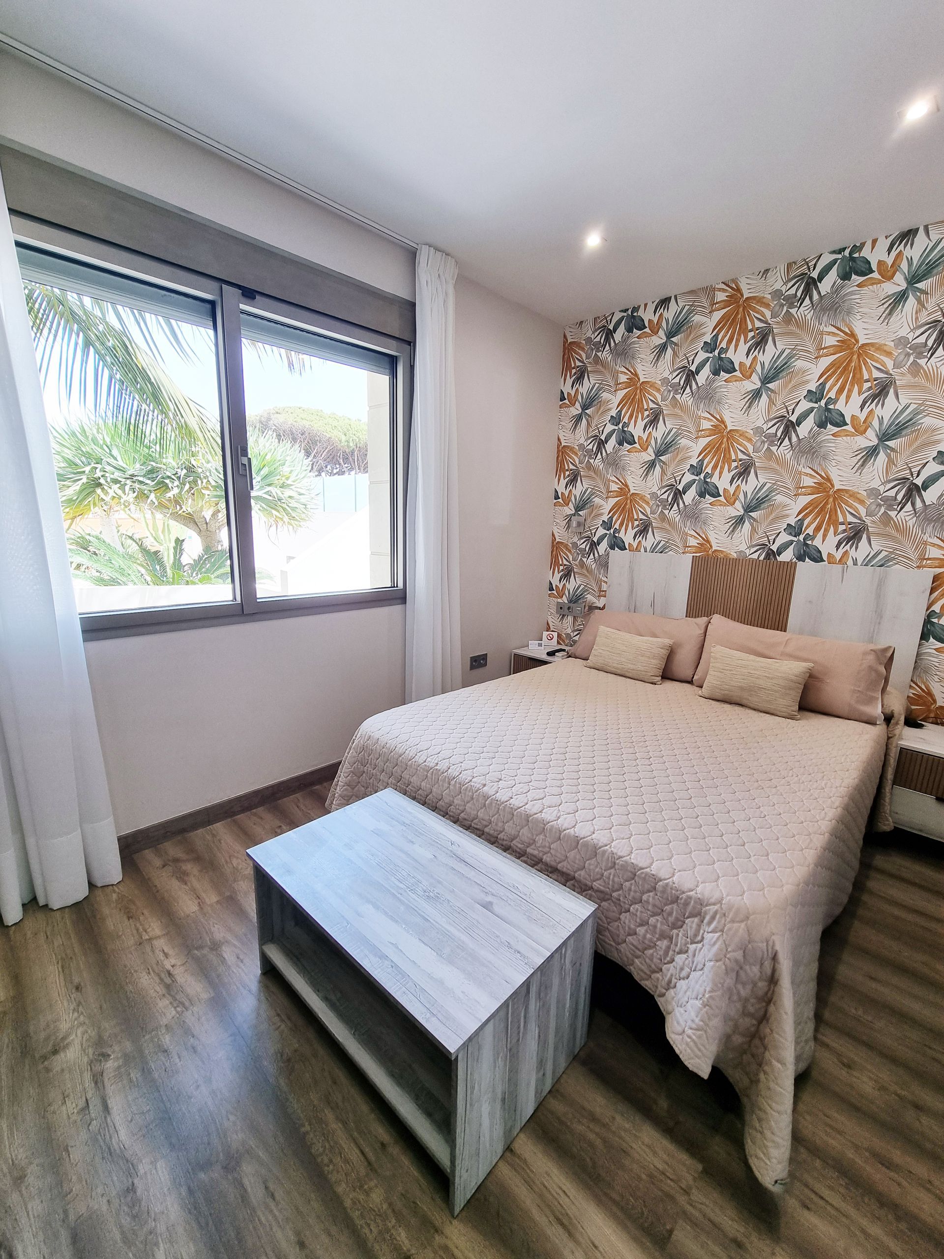 Dormitorio con pared con acento floral, cama con ropa de cama beige, banco y ventana con vista.
